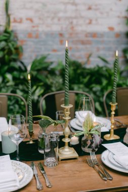 Uzun yeşil mumlar, cam eşyalar ve dekoratif bitkilerle güzel düzenlenmiş bir yemek masası modern bir restoran ortamında misafirperver bir ortam yaratıyor..
