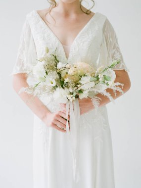 Bir gelin, beyaz ve pastel çiçeklerle dolu yumuşak bir buket tutan, dantel detaylarla dolu güzel beyaz bir elbisenin içinde zarif bir şekilde duruyor. Arkaplan basit ama zarif..