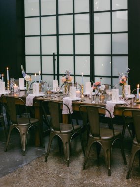 Güzel, uzun, ahşap bir masa narin çiçek aranjmanları ve mumlar sergiliyor ve şık bir yerde akşam toplantısı için sıcak bir ortam yaratıyor..