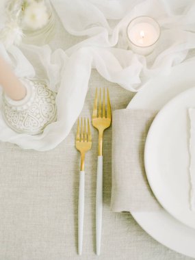 Güzel düzenlenmiş bir yemek masasında altın aksanlı çatallar, yumuşak keten bir peçete ve yumuşak bir mum ışığı sergilenir. Huzurlu ve davetkar bir atmosfer yaratır..