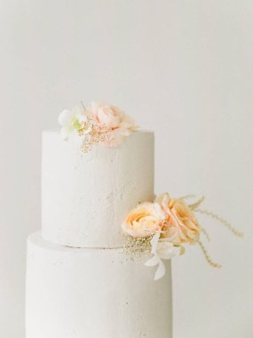 İki katlı beyaz pasta, yumuşak bir zemin üzerinde zarif bir şekilde duruyor. Zarif pastel çiçeklerle süslenmiş. Bu pasta düğünler ya da özel günler için mükemmel. Basit güzellik ve çekicilik sergiliyor..