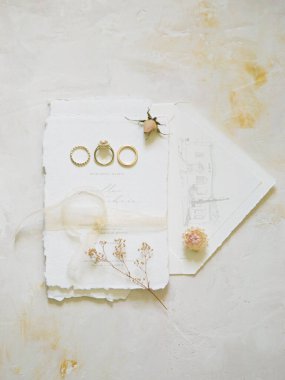 Üç altın alyans, narin çiçekler ve dokulu beyaz kağıt üzerinde zarif bir kurdele içeren güzel bir düzenleme. Düğün için mükemmel..