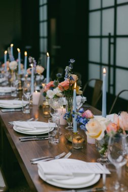 Pastel çiçeklerin, mavi mumların ve zarif sofra takımlarının olduğu güzel bir yemek masası. Dekor, şık ve çağdaş bir mekanda özel bir kutlama için tasarlandı..