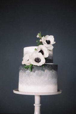 Büyüleyici iki kademeli bir pasta, koyu renkli gerçekçi beyaz çiçeklerle süslenmiş pürüzsüz gri bir ombre tasarımı sunar. Basit bir beyaz duruşta koyu bir arka plana karşı zarif bir şekilde sergilenir..