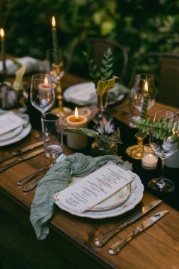 Güzel düzenlenmiş bir açık hava yemek masası şık bulaşık takımları, titreyen mumlar ve basılı menüler sergiliyor. Yeşillik, çevreye romantik bir dokunuş katar..
