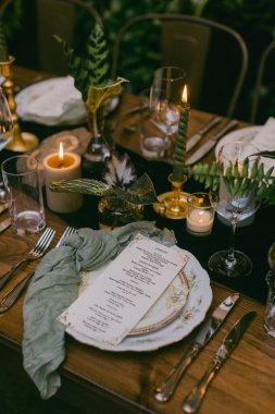 Güzel düzenlenmiş bir masada hassas tabaklar, basılı bir menü ve titreyen mumlar sergileniyor. Yıldızların altında sıcak bir atmosfer yaratıyor..