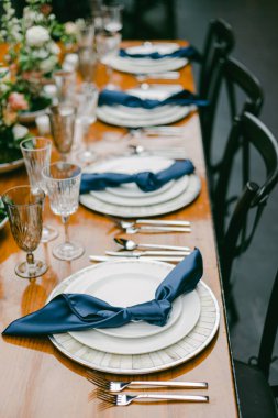 Uzun ahşap bir masa kusursuz bir şekilde tabaklar, cam eşyalar ve cilalanmış çatal bıçaklarla döşenmiştir. Mavi peçeteler özel bir durum için zarif yemek düzenine incelik katar..