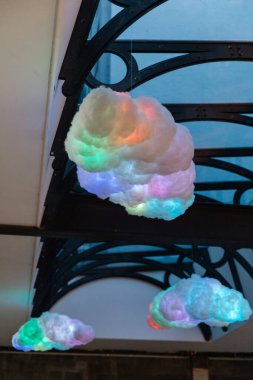 Yumuşak malzemelerden oluşan parlak renkli bulutlar kapalı alanda tasarlanmış karmaşık bir metal kemerden sarkıyor ve tuhaf bir atmosfer yaratıyor..