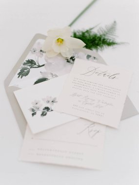 Güzel bir düğün davetiyesi yumuşak bir yüzeyde sergilenir. Davetiyelerde çiçekli tasarımlar ve zarif tipografi yer alıyor, yanında da narin beyaz bir çiçek ve yeşillik var..