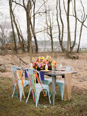 Güzel bir açık hava masasında canlı bir çiçek süsü ve sakin bir gölün yanında renkli sandalyeler yer alır. Sahne ilkbaharın özünü çıplak ağaçlarla yakalıyor..