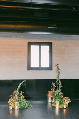 Davetkar bir iç mekanda, büyük bir pencere çerçeveli, yeşillik, bitkiler ve yumuşak mum ışığıyla dikkatlice düzenlenmiş iki çiçek sergisi sergileniyor..