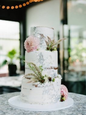 Büyüleyici üç katlı düğün pastası zarif beyaz süslemeler, narin çiçekler ve yeşillikler içerir. Hepsi de hoş, romantik bir ortamda, yumuşak ışıklandırmayla sergilenir..