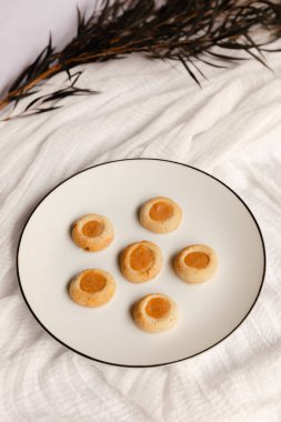 Altın kayısı merkezli altı küçük kurabiye güzel bir şekilde beyaz bir tabağa konur. Kırsal alan yumuşak kumaşlar ve biraz yeşillik içeriyor, rahatlatıcı bir atmosfer yaratıyor..