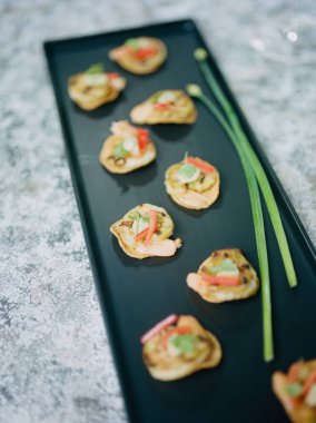 Süslü küçük, aromalı ısırıklar siyah bir tabakta güzelce sunuluyor. Taze malzemeler etkinlikte davetkar bir atmosfer yaratıyor, mutfak sanatı sergileniyor..