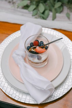 Şık bir masa düzeninde kremalı katmanlarla dolu ve üzeri çilek, ahududu ve böğürtlen dolu bir tatlı bardağı zarif bir şekilde peçeteyle sunuluyor..
