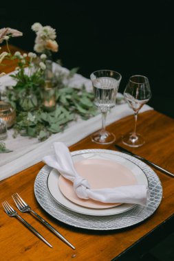 Güzel düzenlenmiş bir masa güzel sofra eşyaları, cam eşyalar ve çiçekli bir süs eşyası içerir. Samimi bir yemek deneyimi için ortam mükemmel, sıcak bir atmosfer yaratıyor..