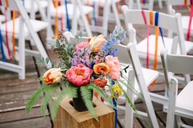 A vibrant floral centerpiece features peonies and delphiniums, set on wood near rows of chairs with colorful ribbons. clipart