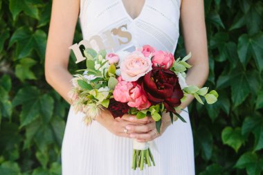 Bir gelin, üzerinde pembe güller ve şakayıklar olan canlı bir buket tutar. Yanında da yeşil yaprakların arasında duran bir bayan işareti..