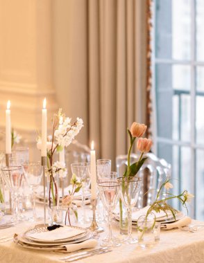 Cam eşyalar, çiçekler ve mumlarla güzel düzenlenmiş yemek masası misafirler için sıcak bir ortam yaratıyor..