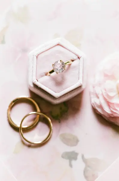 Göz kamaştırıcı bir elmas nişan yüzüğü iki altın alyans ve yumuşak çiçeklerle çevrili narin bir kutunun üzerinde duruyor..