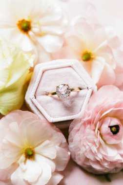 Göz kamaştırıcı bir elmas yüzük zarif pastel çiçeklerle çevrili yumuşak bir kadife kutunun üzerinde duruyor ve romantik bir ortam yaratıyor..