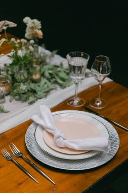 Güzel düzenlenmiş bir masa güzel sofra eşyaları, cam eşyalar ve çiçekli bir süs eşyası içerir. Samimi bir yemek deneyimi için ortam mükemmel, sıcak bir atmosfer yaratıyor..