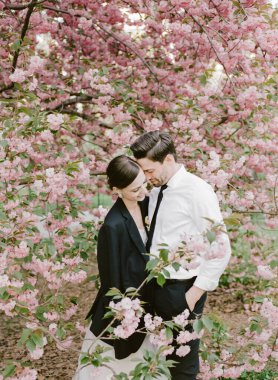 Resmi giyinmiş bir çift, canlı pembe kiraz çiçekleriyle çevrili samimi bir anı paylaşıyor..