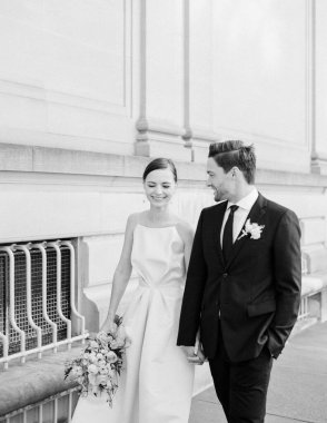 Düğün kıyafetleri içinde neşeli bir çift, tebessümlerini paylaşıyorlar ve arkalarında şehir mimarisi ile bir buket tutuyorlar..