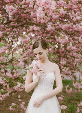 Üzerinde straplez bir elbise olan gelin, elinde bir buket, kiraz çiçeklerinin arasında duruyor, sakin bir bahar öğleden sonrası sergiliyor.
