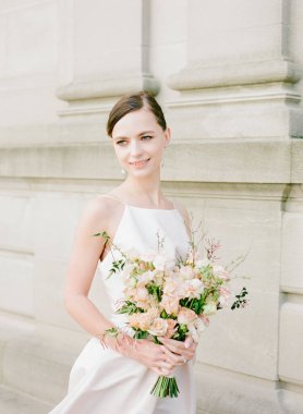 Gülümseyen bir gelin zarif bir şekilde elinde büyüleyici bir çiçek buketini tutarak tarihi bir mekânın cazibesinin tadını çıkarıyor..