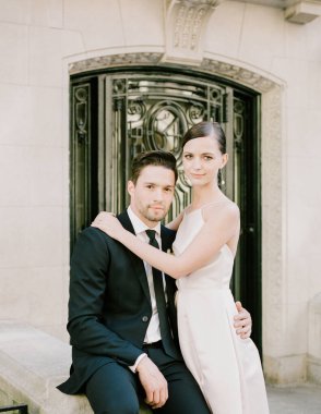 Resmi giyinmiş bir çift, taş bir çıkıntıda yan yana oturur, dekoratif bir kapının önünde samimi bir anı paylaşırlar..