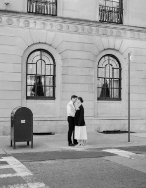 Bir çift, gün ışığıyla dolu kentsel bir ortamda, klasik binaların yanında kucaklaşarak, hassas bir anı paylaşıyor..