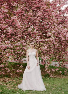Bir gelin, güneşli bir bahar gününde parlak pembe kiraz çiçeklerinin önünde beyaz bir elbiseyle güzel poz verir..