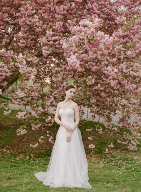 Göz kamaştırıcı beyaz elbiseli bir gelin güneşli bir bahar gününde canlı pembe kiraz çiçeklerinin arasında poz verir..