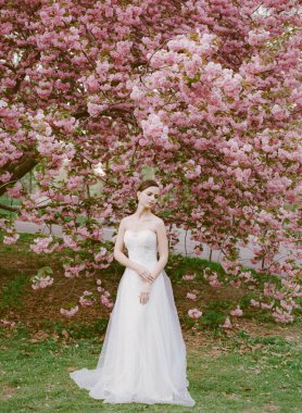 Bir gelin canlı pembe kiraz çiçeklerinin altında zarif bir şekilde poz verir, güneşli bir bahar gününde yemyeşil ile çevrili..