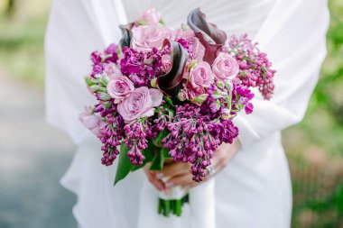 Bir gelin güzel bir demet pembe ve mor çiçek sergiliyor manzaralı bir arka planda.