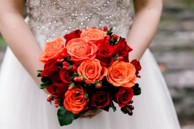 Narin elbiseli bir gelin, yemyeşil bir bahçede canlı bir buket turuncu ve kırmızı gül tutar..