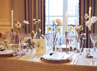 Çok güzel düzenlenmiş bir yemek masasında narin çiçekler, mumlar ve hoş bir ortamda ince cam eşyalar bulunur..