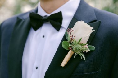 Siyah smokin giymiş şık bir damat, açık havada romantik bir düğün sırasında beyaz bir gül yaka çiçeğini sunar..