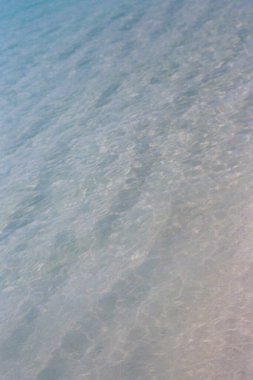 Yumuşak dalgalar kıyıya vuruyor, güneş ışığı berrak bir sahilde parıldıyor..