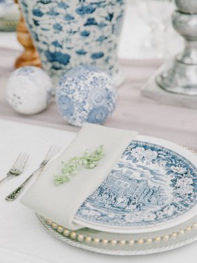 Bir yemek masasında güzel mavi ve beyaz porselen tabaklar, zarif gümüş çatallar ve dekoratif çiçek düzenlemeleri sergilenir..