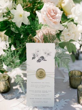 Çok güzel bir düğün davetiyesi pastel güllerin ve zambakların arasında duruyor. Parlayan mum tutucularla birlikte. Kutlama için büyüleyici bir atmosfer yaratıyorlar..