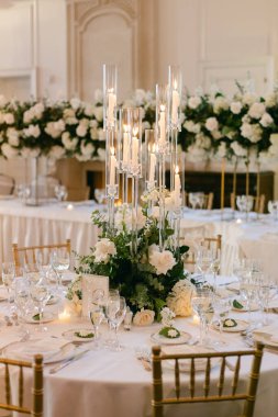 Zarif yemek masasında beyaz çiçekler, mumlar ve cam eşyalar var. Bir ziyafet salonunda özel bir etkinlik için hazırlanıyorlar..