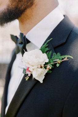 Keskin siyah takım elbiseli bir damat beyaz bir çiçek yaka çiçeği sergiliyor. Doğal yeşillik, bulanık bir arka plana karşı zarif çiçek düzenini tamamlıyor..