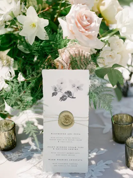 Çok güzel bir düğün davetiyesi pastel güllerin ve zambakların arasında duruyor. Parlayan mum tutucularla birlikte. Kutlama için büyüleyici bir atmosfer yaratıyorlar..