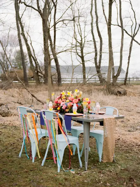 Güzel bir açık hava masasında canlı bir çiçek süsü ve sakin bir gölün yanında renkli sandalyeler yer alır. Sahne ilkbaharın özünü çıplak ağaçlarla yakalıyor..