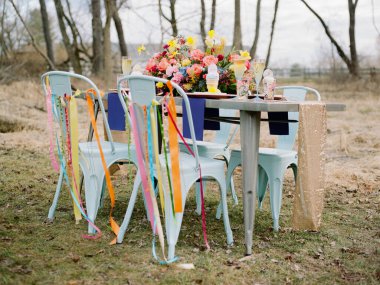 Büyüleyici bir açık hava masası, canlı bir çiçek süsü, renkli sandalyeler ve sakin doğal bir fon ile süslenmiş bir bahar kutlaması için ayarlandı..