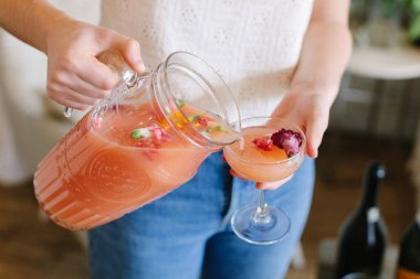 Bir insan taze çiçekler ve yapraklarla çevriliyken sürahiden zarif bir bardağa canlı bir kokteyl döker..