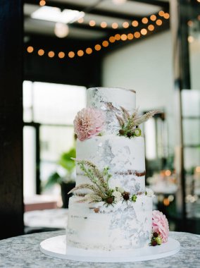 Çok güzel tasarlanmış bir düğün pastası üç katlı, yumuşak pembe çiçekler ve yeşilliklerle süslenmiş, bir resepsiyon salonunda bir arkaplan dolusu sıcak ışığın karşısında duruyor..
