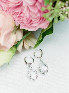 Bir buket yumuşak pembe çiçek ve yeşil yaprakların yanında duran kristalli zarif damla küpeler..